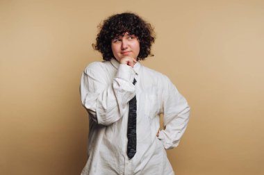 A person stands confidently in a white shirt with rolled-up sleeves and a black tie. They have curly hair and a thoughtful expression, as they ponder something interesting.