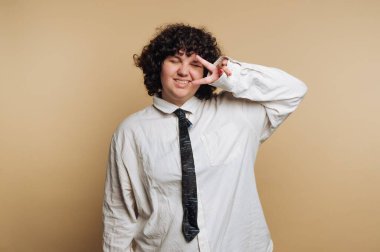 A person with curly hair smiles and makes a peace sign while wearing a loose white shirt and a black tie. The setting features a simple beige background, creating a cheerful atmosphere.