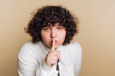 A young person with curly hair is posing with a finger to their lips, signaling for silence. The backdrop is a warm, neutral color, creating a calm atmosphere.