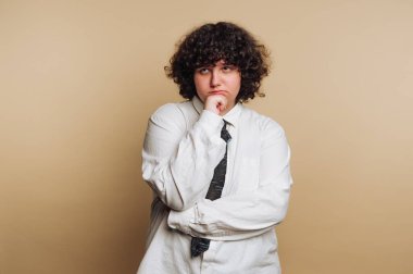 A person with curly hair stands thoughtfully with one hand on their chin. They wear a white shirt and a tie, against a plain beige background. The expression reflects deep thought.