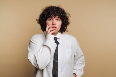 A person with curly hair stands in front of a light beige backdrop, dressed in a large white shirt and a black tie. They appear thoughtful, with a hand on their cheek.