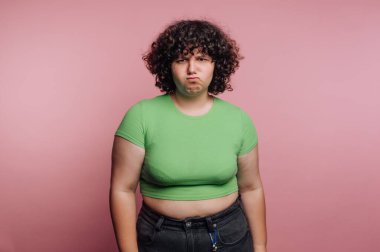 A young person with curly hair poses in front of a pink backdrop. They wear a bright green shirt and sport a playful expression, adding a fun vibe to the scene.