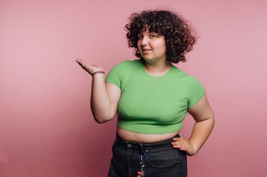 A young woman stands confidently with her hand raised in a casual gesture. She wears a green fitted shirt and black jeans, set against a soft pink backdrop. Her curly hair adds charm to the scene.