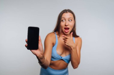 Enthusiastic woman points at her smartphone with a surprised smile in a bright studio setting. She is dressed casually and appears excited about something on the screen.