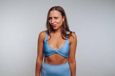 A woman stands confidently in a blue top and matching bottom, playfully expressing her emotions in a minimalist studio. The background is neutral, emphasizing her charm.