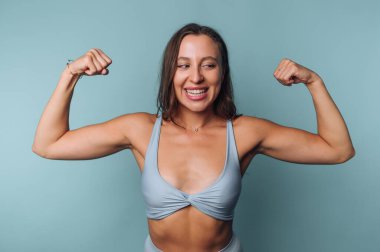 A confident woman shows her strength by flexing her arms and smiling. She wears a light blue athletic outfit and stands in front of a solid blue backdrop, radiating positivity and empowerment.