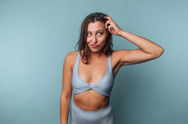 A woman displays a playful expression while scratching her head in a bright blue studio. She wears a light blue outfit and has long, wavy hair, adding to the lively atmosphere of the scene.