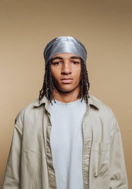 A young person stands with confidence, wearing a light-colored shirt and a silver headwrap. The neutral background highlights the relaxed, modern style and individuality of urban fashion.