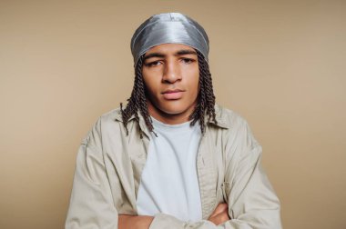 A young man stands with arms crossed, displaying a confident expression. He wears a light shirt and a silver headscarf, set against a beige backdrop that highlights his features and style.