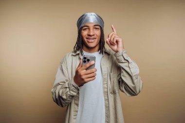 A young man with curly hair wearing a silver headscarf and beige shirt points upward with a smile while holding a smartphone in a warm indoor setting.