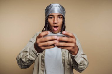 A young man with braided hair and a silver head wrap holds a gaming console, revealing a surprised expression as he reacts to a gaming moment. The indoor setting adds focus to his excitement.