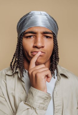 A young man stands against a light backdrop, wearing a silver head wrap. He has braided hair and a contemplative expression, resting his finger on his lips as if deep in thought.