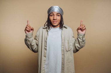 A person stands confidently with both hands raised, fingers pointing upwards, dressed in a casual outfit with a long shirt. The background is simple and warm-toned.