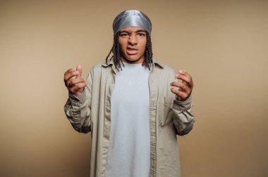 A young person stands against a plain background with a look of frustration. They wear a casual outfit and a silver head wrap, gesturing with their hands.
