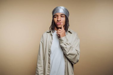 A young man stands with a hand on his chin, deep in thought. He wears a silver headscarf and a light-colored shirt, showcasing a calm expression.