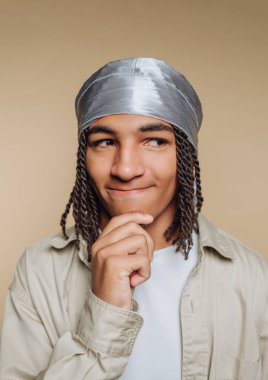 A young man with braided hair is posing thoughtfully, hand on chin, wearing a shiny silver head wrap in front of a neutral background. His expression shows contemplation.