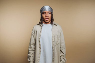 A young individual stands with an astonished look, wearing a light-colored shirt and a silver headscarf. The backdrop is a soft beige color, creating a calm setting.