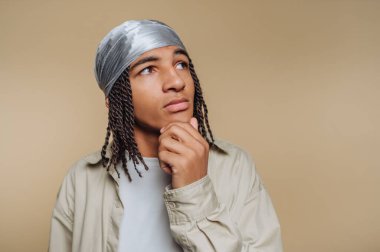 A young person wearing a glossy headscarf poses thoughtfully against a plain background. Their facial expression reflects contemplation and curiosity, inviting viewers to wonder about their thoughts.