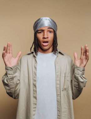 A young man stands with raised hands, wearing a silver headscarf and a casual shirt. His facial expression shows surprise or confusion, set against a plain background.