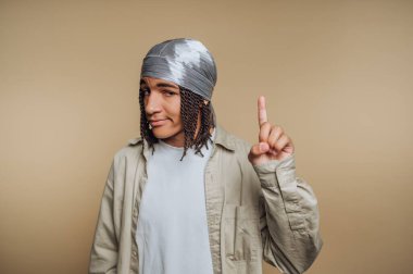 A young man stands against a light brown wall, wearing a gray bandana and a loose white shirt. He points up with a playful expression, showcasing his unique style and confidence.