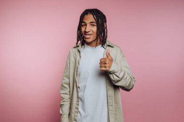 A young individual stands confidently against a pink backdrop, smiling and giving a thumbs up. They wear a casual outfit that adds to the relaxed vibe of the moment.