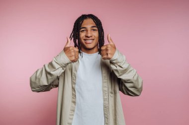 A young man with braided hair gives a thumbs-up in front of a soft pink background. He is wearing a casual shirt and has a cheerful expression on his face.