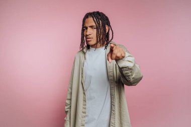 A young man stands confidently with a serious look, pointing directly at the viewer against a solid pink background. He wears a casual outfit that adds to the relaxed vibe of the scene.