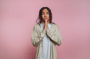 A young individual stands with hands together in a gesture of hope. The soft pink background enhances the emotion of the moment, showcasing a sense of vulnerability.
