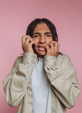 A young man with braided hair stands against a soft pink background, exhibiting a fearful expression as he grips his face with his hands. His casual outfit adds to the relaxed atmosphere.