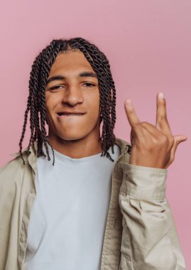 A young man with braided hair poses confidently, flashing a cheerful hand sign. His casual outfit and bright expression stand out against the pink backdrop.
