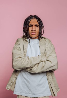 A young man stands with crossed arms, showcasing a serious expression. He is dressed casually in a light-colored shirt and jacket, with a pink backdrop enhancing the focus on him.