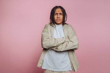 A young man stands with crossed arms and a serious expression, wearing a light-colored outfit. The plain pink background enhances his confident demeanor, creating a striking visual.