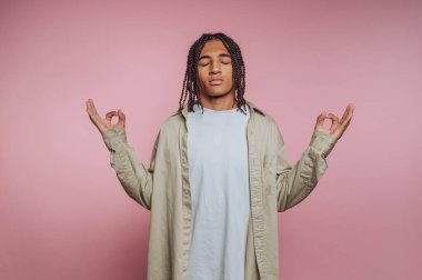 A person with braided hair is meditating calmly in a serene pose, wearing a loose, light-colored shirt. The soft pink background enhances a peaceful atmosphere aimed at relaxation.