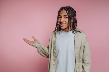 A young individual wearing a casual outfit stands against a soft pink backdrop. They smile and use hand gestures to convey a friendly and approachable attitude.
