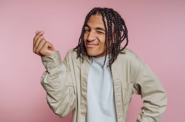 A young man shows a playful expression while winking and using his hand to make a gesture. He stands against a soft pink background, highlighting his relaxed style and confidence.