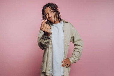 A young individual with braided hair playfully poses, making a heart shape with their fingers in front of a soft pink backdrop. The relaxed outfit adds to the cheerful vibe.