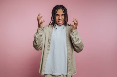 A young individual with braided hair shows a frustrated expression, hands raised in frustration, wearing a light shirt and cream-colored pants in a cozy indoor setting.