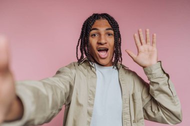 A cheerful young man shows excitement while waving his hand in greeting. He stands in front of a soft pink backdrop, wearing a light shirt and a casual jacket, creating a lively atmosphere.