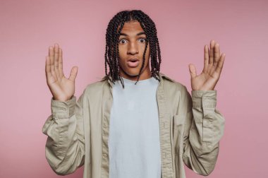 A young man with braided hair stands against a pink backdrop, raising his hands in surprise. His expression is playful and engaging, capturing a lighthearted moment.