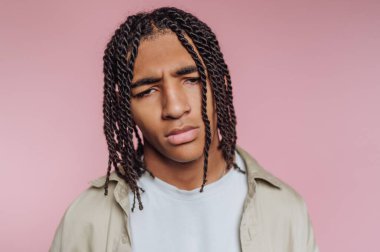 A young man with braided hair stands confidently in front of a soft pink background. He displays a thoughtful expression while wearing a light shirt. This moment captures a sense of calm.