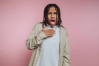 A young individual stands against a pink backdrop, displaying an expression of surprise with their mouth open and hand on their chest. The casual attire adds a relaxed vibe to the scene.