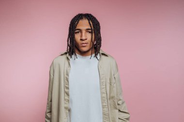 A young man with braided hair stands confidently against a soft pink background, wearing a casual outfit. He gazes directly at the camera, showcasing a relaxed yet strong expression.