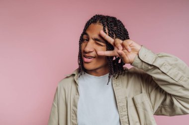 A young man smiles while making a peace sign with his fingers. He has braided hair and wears a light-colored shirt, standing in front of a pink wall.