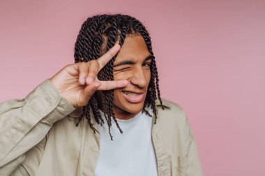 A young individual shows a cheerful expression while making a peace sign with one hand. The playful pose is set against a soft pink backdrop, creating a lively atmosphere.