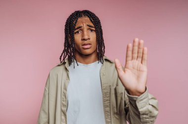 A young man with braided hair stands with a worried expression, raising his hand in a gesture that suggests stopping or caution against something. The soft pink background adds a calming tone.