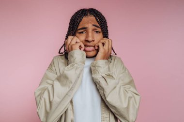 A young individual displays feelings of anxiety while in a studio. The background is pink, and the person is holding their face, expressing nervousness during the shoot.
