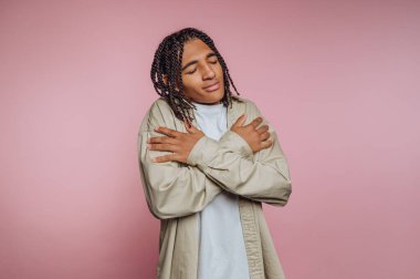 A young individual stands in a studio with a light pink backdrop, embracing themselves with a smile. Their relaxed pose conveys a sense of self-acceptance and tranquility.