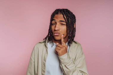 A young man with braided hair is posing in front of a soft pink background. He has a thoughtful expression, with one finger on his chin and a playful wink.