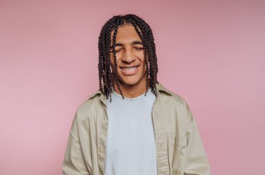 A young individual with braided hair and braces is smiling happily. They are wearing a light-colored shirt and standing in front of a pink backdrop, showing a relaxed and cheerful mood.