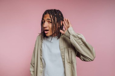 A young man with braided hair is leaning in with a playful expression, as if he is listening intently. The background is a soft pink, creating a vibrant atmosphere.